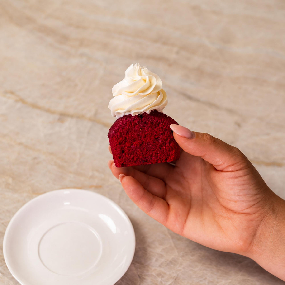 Hand holding a vibrant red cupcake topped with white frosting above a small white saucer on a marble surface.
