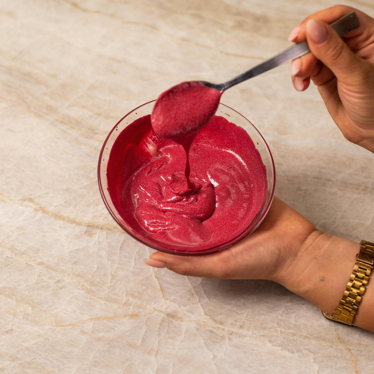 A hand holding a glass bowl of vibrant red food coloring paste, with a metal spoon drizzling the mixture.