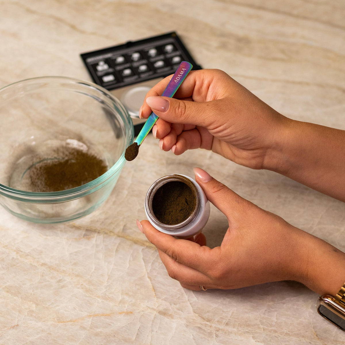 Gray natural food coloring powder being added to glass bowl with a measuring spoon.