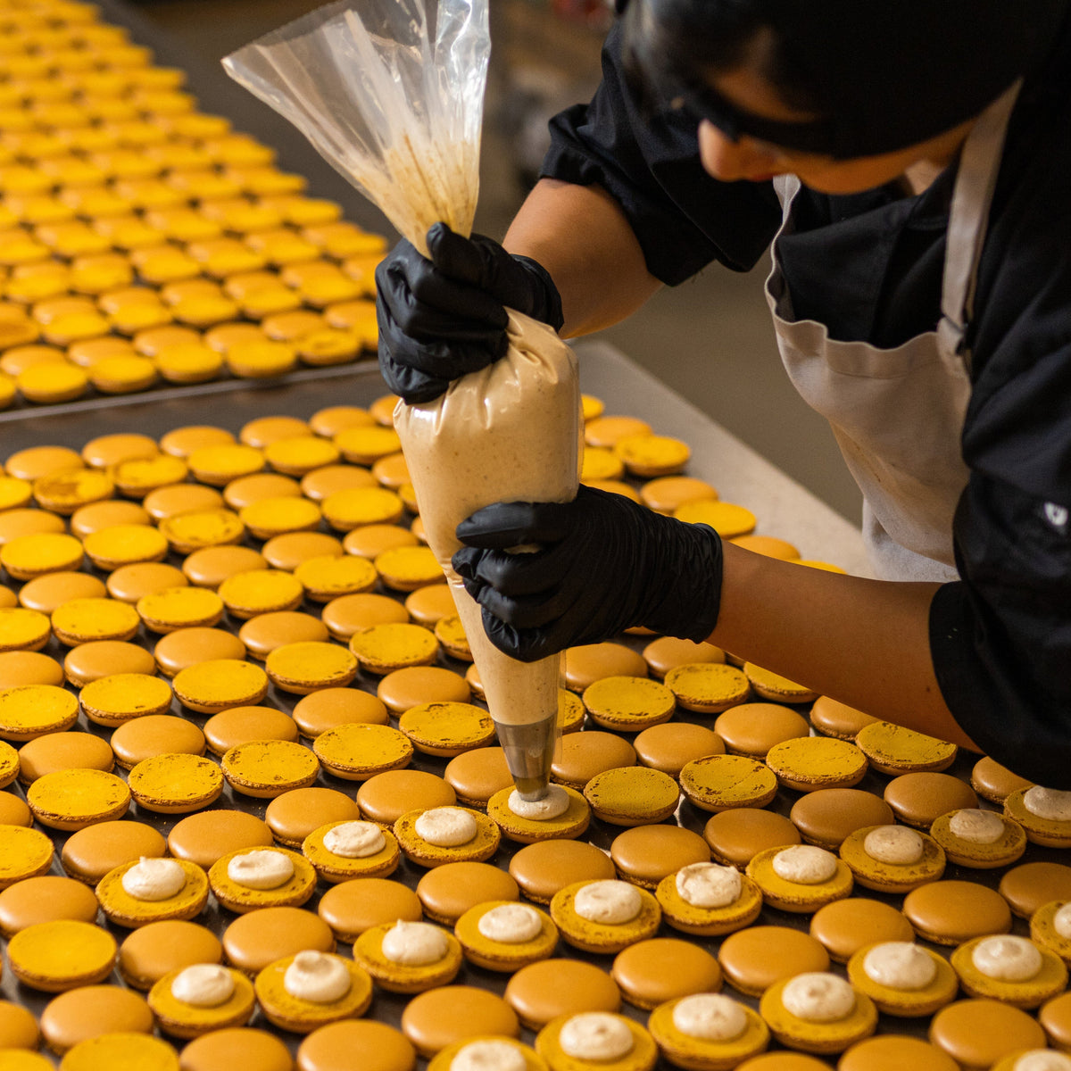 Baker piping pumpkin filling onto rows of vibrant orange pumpkin macaron shells 